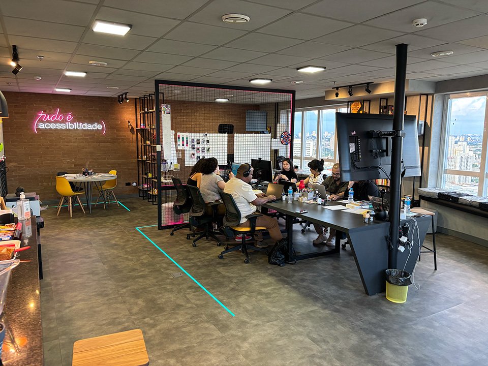 Um ambiente de escritório moderno e colaborativo. No centro, o grupo trabalha em uma mesa grande com notebooks e materiais diversos. À esquerda, há uma parede com um letreiro em neon rosa que diz “tudo é (acessibilidade)”. O espaço é bem iluminado, possui janelas amplas com vista para a cidade e decoração contemporânea com móveis coloridos e elementos industriais. Há uma atmosfera criativa e inclusiva.