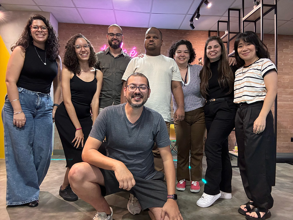 Grupo de oito pessoas que participaram do treinamento posando para a foto, há uma parede de tijolos ao fundo e iluminação de teto. Todos estão sorrindo, demonstrando um clima de amizade e trabalho em equipe. Marcelo Sales está agachado à frente, enquanto as demais pessoas estão em pé lado a lado.