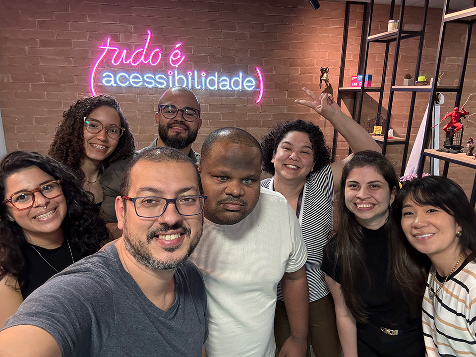 Grupo de oito pessoas sorrindo para uma selfie em um ambiente de escritório moderno. Ao fundo, há uma parede de tijolos com um letreiro em neon que diz “tudo é (acessibilidade)”. As pessoas parecem estar alegres e descontraídas, transmitindo um clima de união e inclusão. Estantes com objetos decorativos também compõem o cenário.