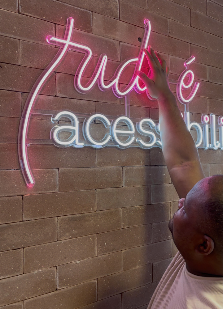 Diego, que é cego e foi um dos participantes do curso, tocando em um letreiro de neon fixado em uma parede de tijolos. O letreiro tem as palavras “tudo é (acessibilidade)” escritas em letras cursivas e iluminadas em rosa e branco. Diego está interagindo com o letreiro de forma sensorial, reforçando simbolicamente a mensagem de inclusão e acessibilidade presente no texto.