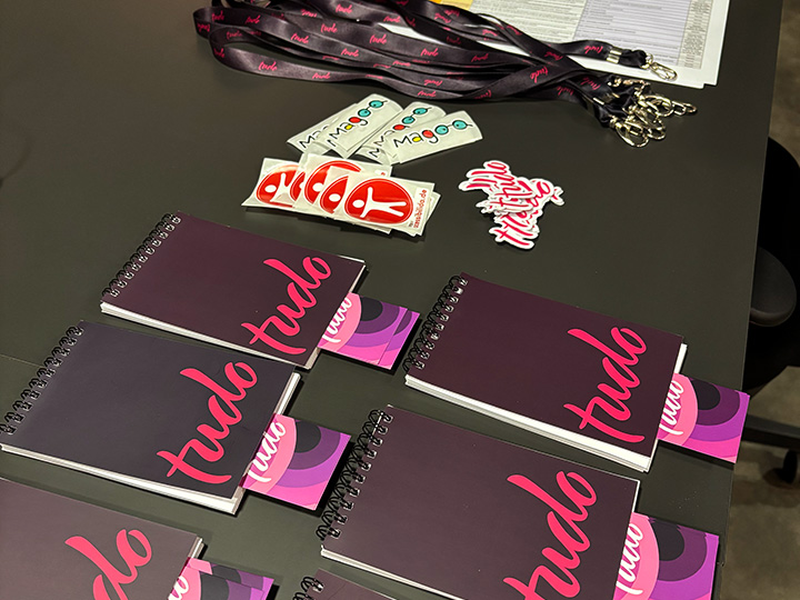 Vários cadernos com a capa roxa e a palavra TUDO em rosa, adesivos coloridos (incluindo logotipos do Magoo e de um personagem vermelho com braços e pernas abertos que representam a acessibilidade), crachás com cordões personalizados e um papel com uma tabela ao fundo. O conjunto sugere um ambiente organizado e criativo, com foco em identidade visual e acessibilidade.